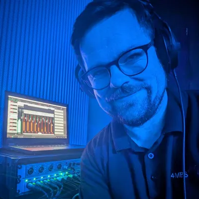 Jai, wearing headphones, smiling in a blue-lit recording booth beside a laptop and a rack of microphone preamps.